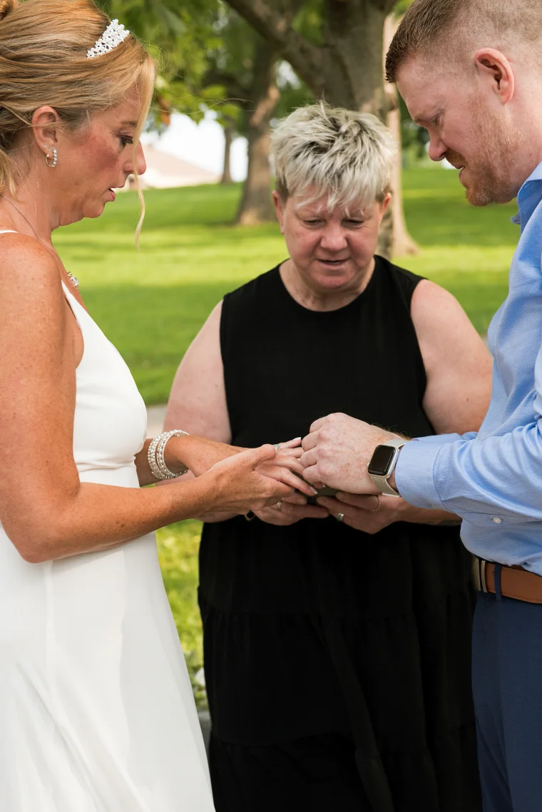 A bride in a pearl headpiece slips a ring onto her finger as the groom guides her hand and the officiant looks on. An intimate outdoor ceremony under shady trees, focused on the moment that matters.
