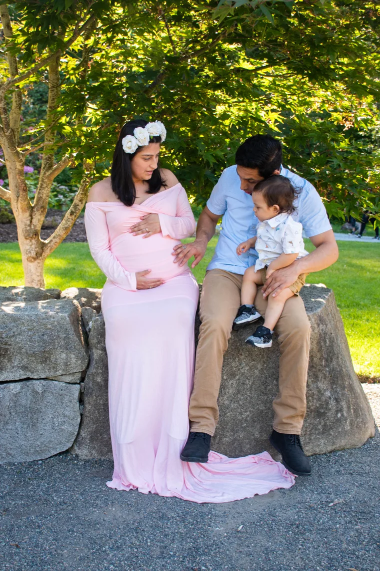 An expecting mother in a flowing blush gown and white rose crown cradles her bump while her partner reaches over, their toddler perched on his lap. Dappled garden light and a stone ledge make a soft, natural setting for a growing family.