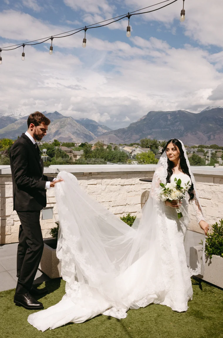 A groom gathers the long lace-trimmed cathedral veil as his bride glances back with a knowing smile. String lights overhead and sweeping mountain views frame the scene.