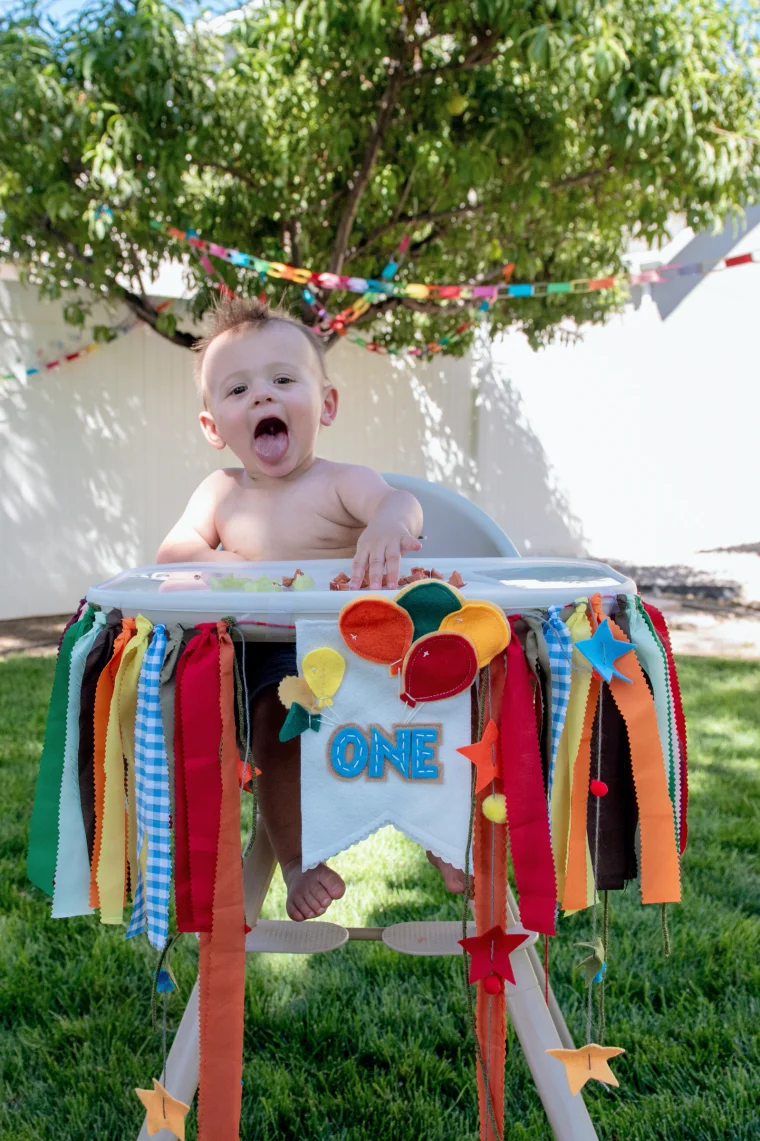 A delighted one-year-old sticks out his tongue from a high chair dressed in a colorful fabric-strip garland and a handmade “ONE” felt banner with balloons. Paper chains strung through the backyard tree finish the celebration.