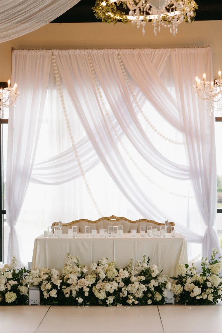 A gold-trimmed velvet settee sits behind a candlelit head table, backed by swagged white drapery threaded with oversized pearl strands. Lush white florals line the base, and crystal chandeliers glow on either side.