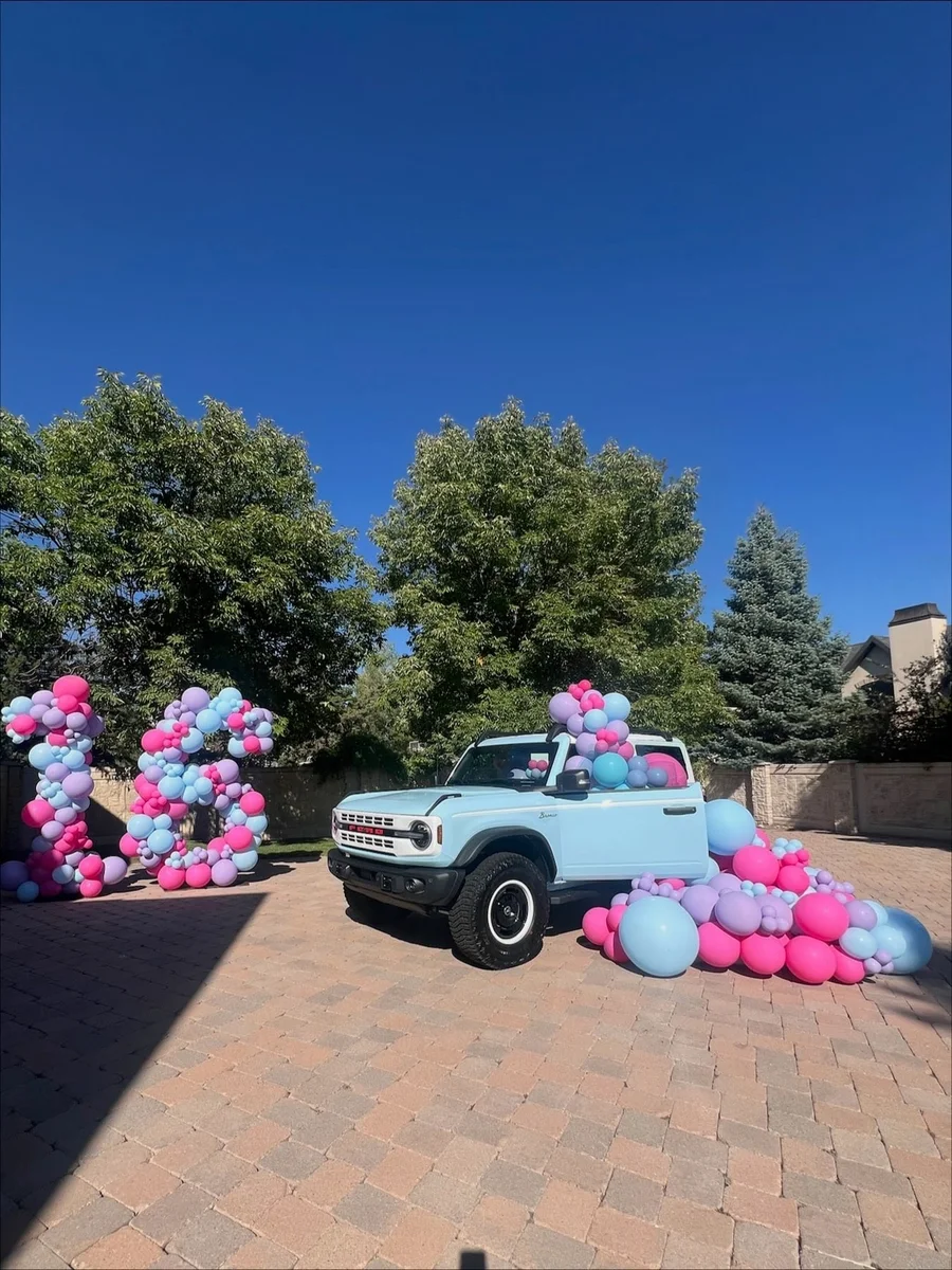 Pastel pink and blue balloon garland around a light blue SUV on a brick driveway