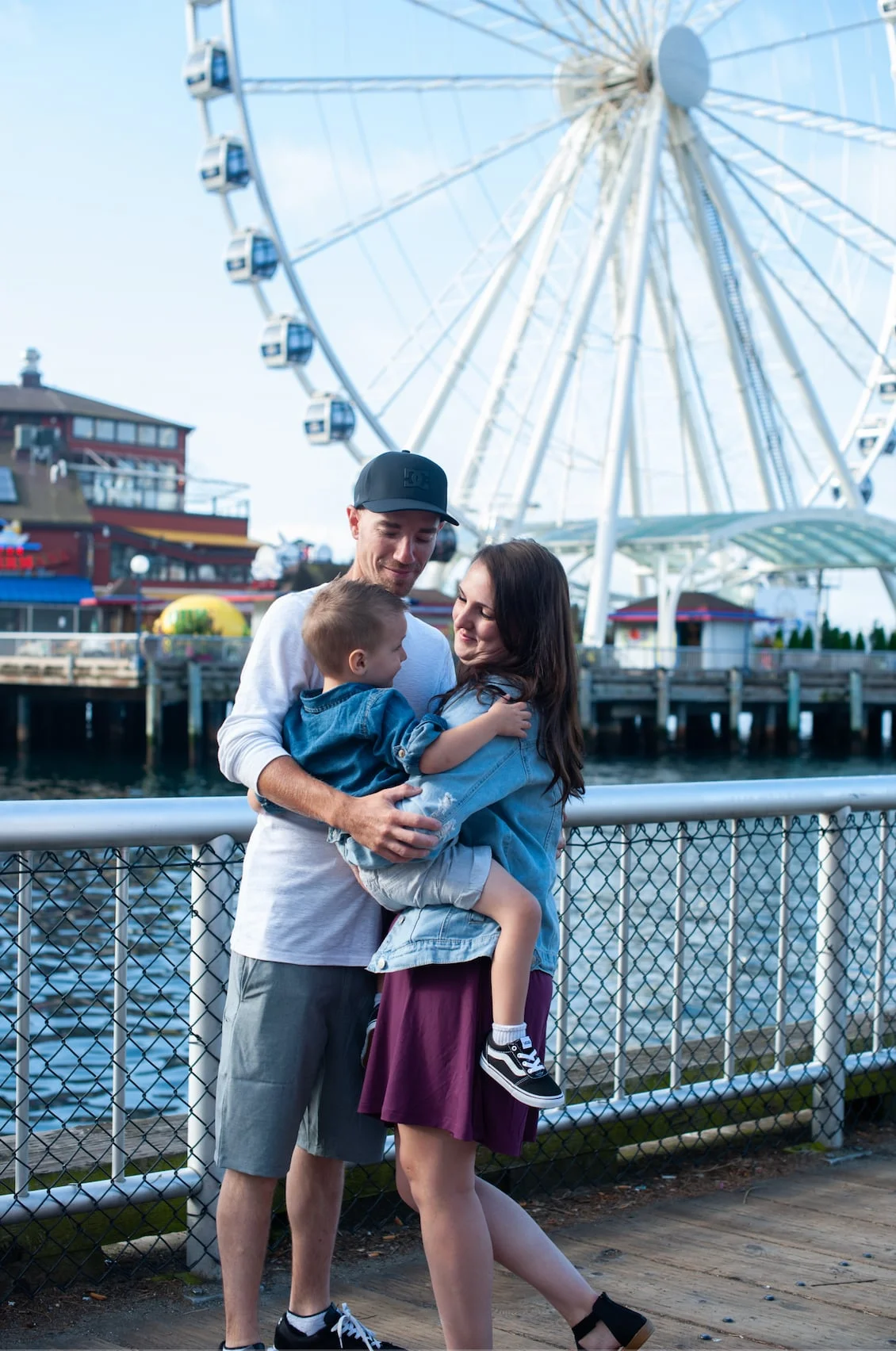 A family of four embracing on a waterfront pier with a Ferris wheel behind them.