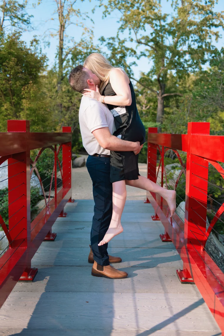 On a bright red footbridge, he lifts her off her feet for a kiss as she cups his face. Leafy trees and late-day light give the moment a playful, cinematic feel.