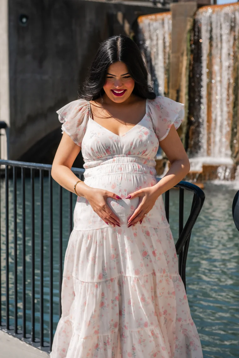 A glowing mom-to-be in a tiered floral chiffon dress forms a heart with her hands over her bump. A cascading fountain sparkles in the background.