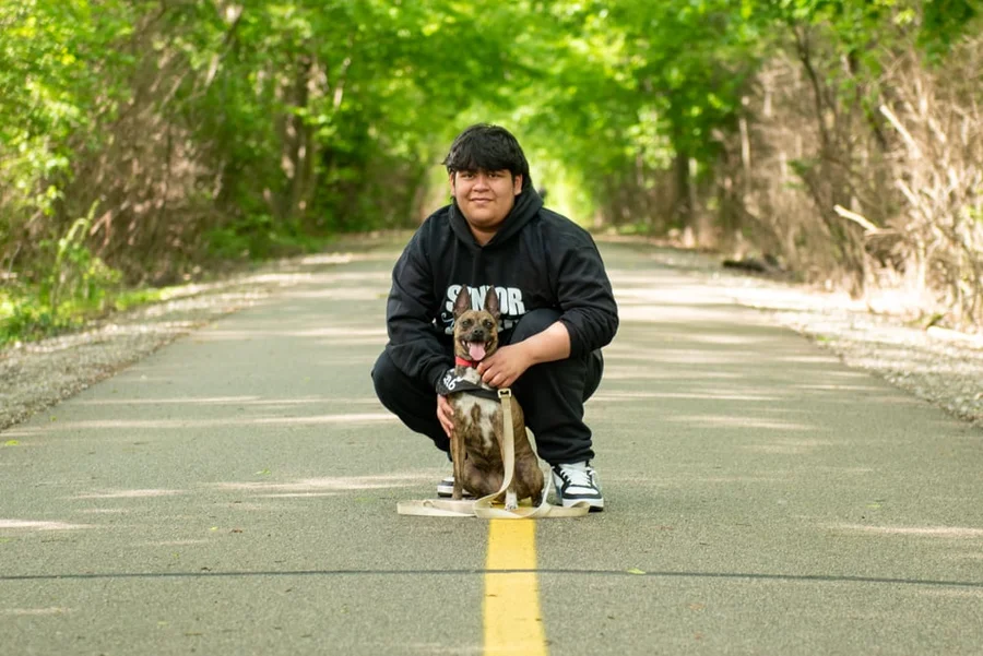 Young person crouching with a dog on a tree-lined paved trail