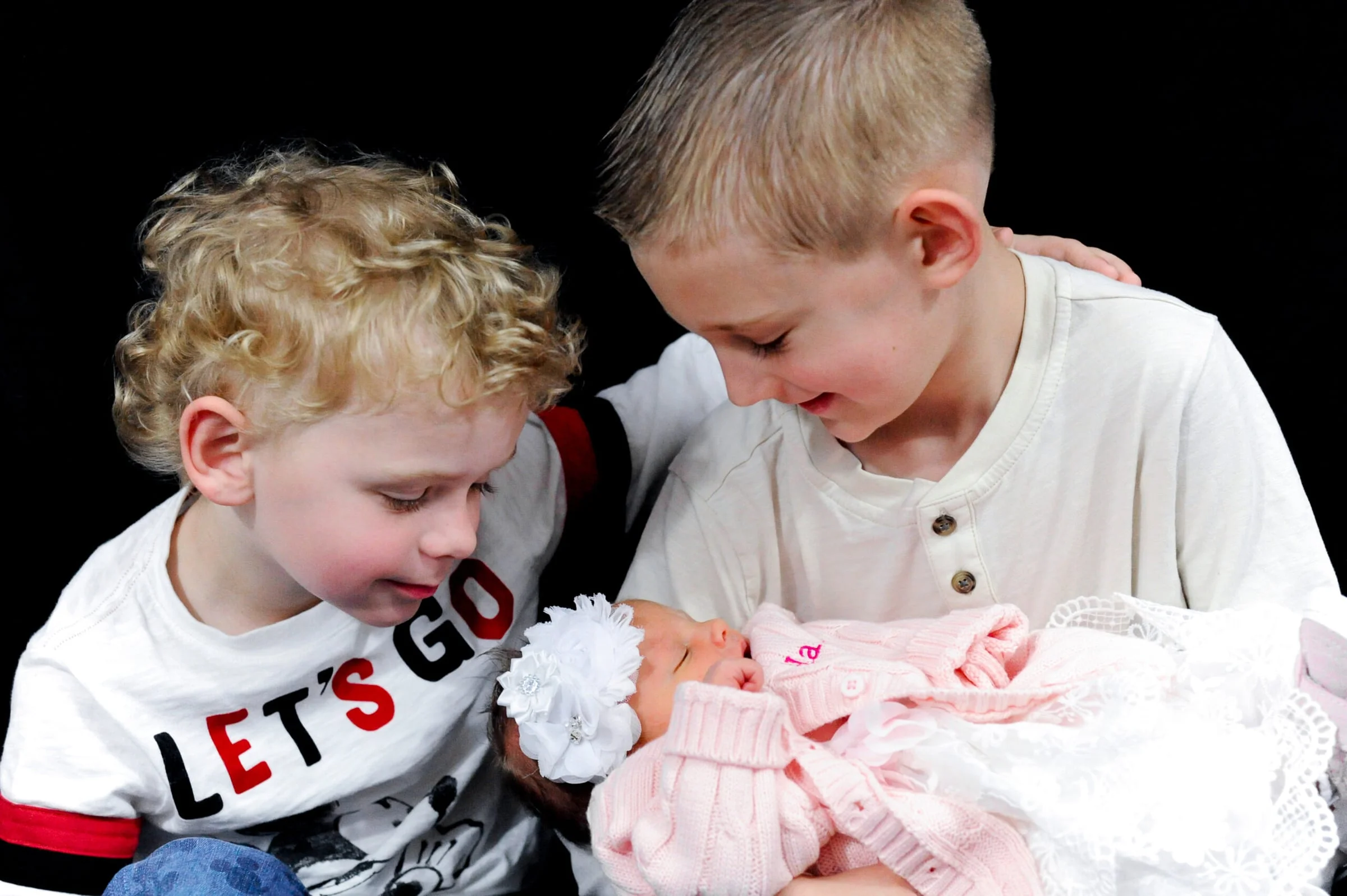 Two young brothers gazing down at their newborn baby sister, who is wrapped in a pink blanket.