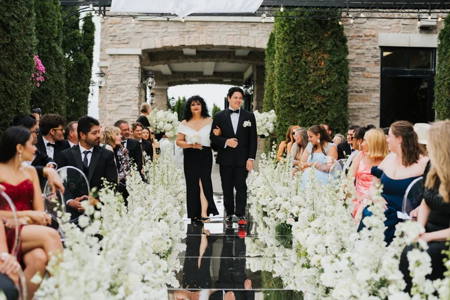 Couple walking down an aisle lined with white flowers toward seated guests