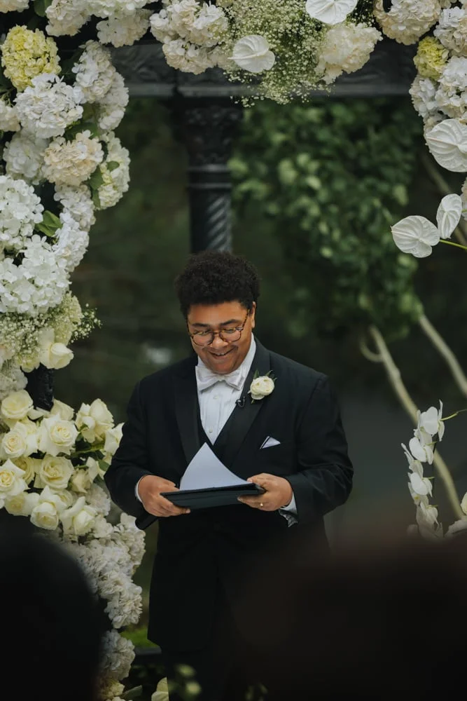 Officiant in a tuxedo reading from a folder before a white floral arch