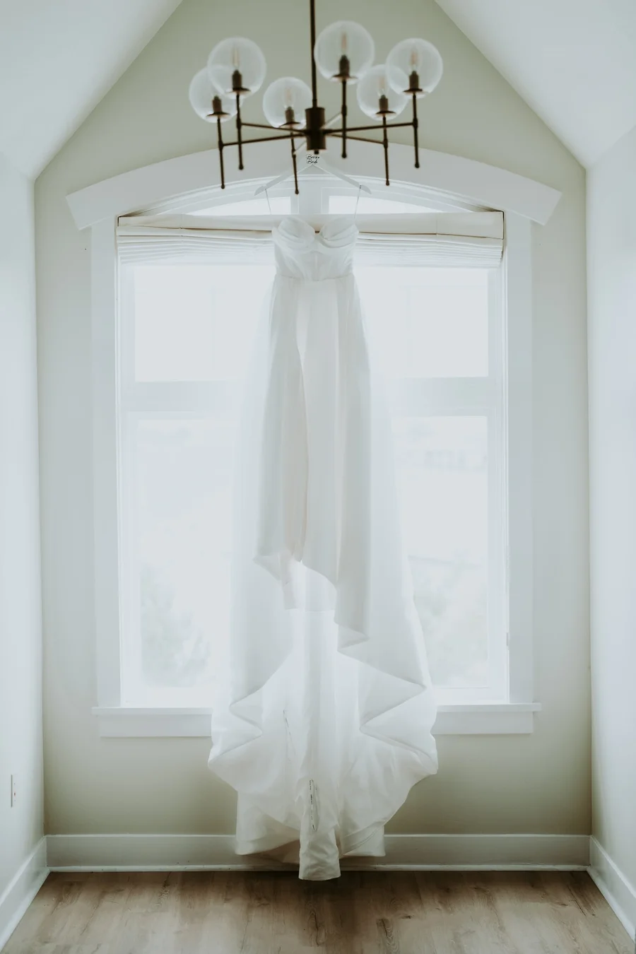 Bridal gown hanging from a chandelier in front of a tall arched window